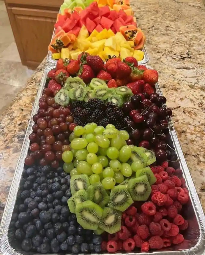 a long aluminum tray filled with neatly arranged, chopped fruits forming a rainbow pattern from top to bottom: watermelon…