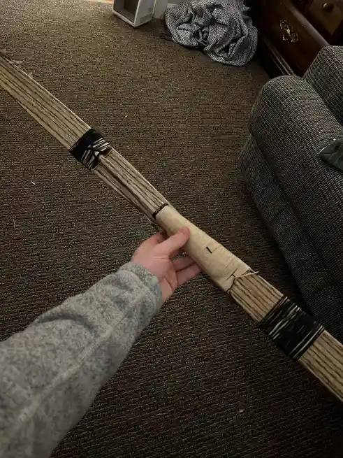 A person's hand holds a long, handmade wooden object resembling a bow or staff, wrapped with black and beige cord, over a…