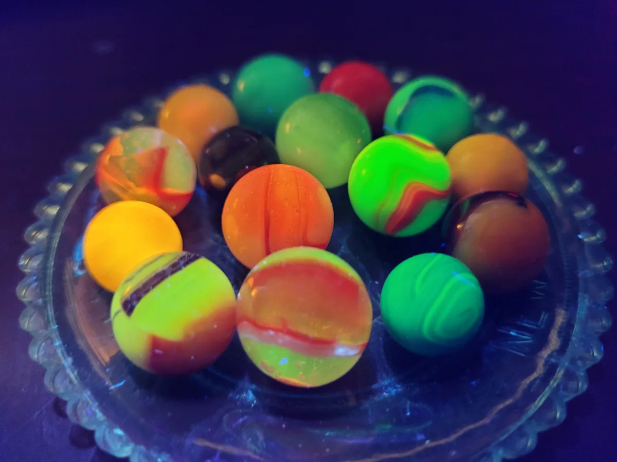 A close-up photo shows a collection of colorful marbles glowing brightly under blacklight in a clear glass dish with a…