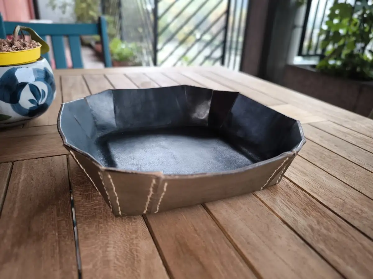 A handmade, dark gray, octagonal leather-like tray with visible stitching sits on a wooden table outdoors. To the left, a…