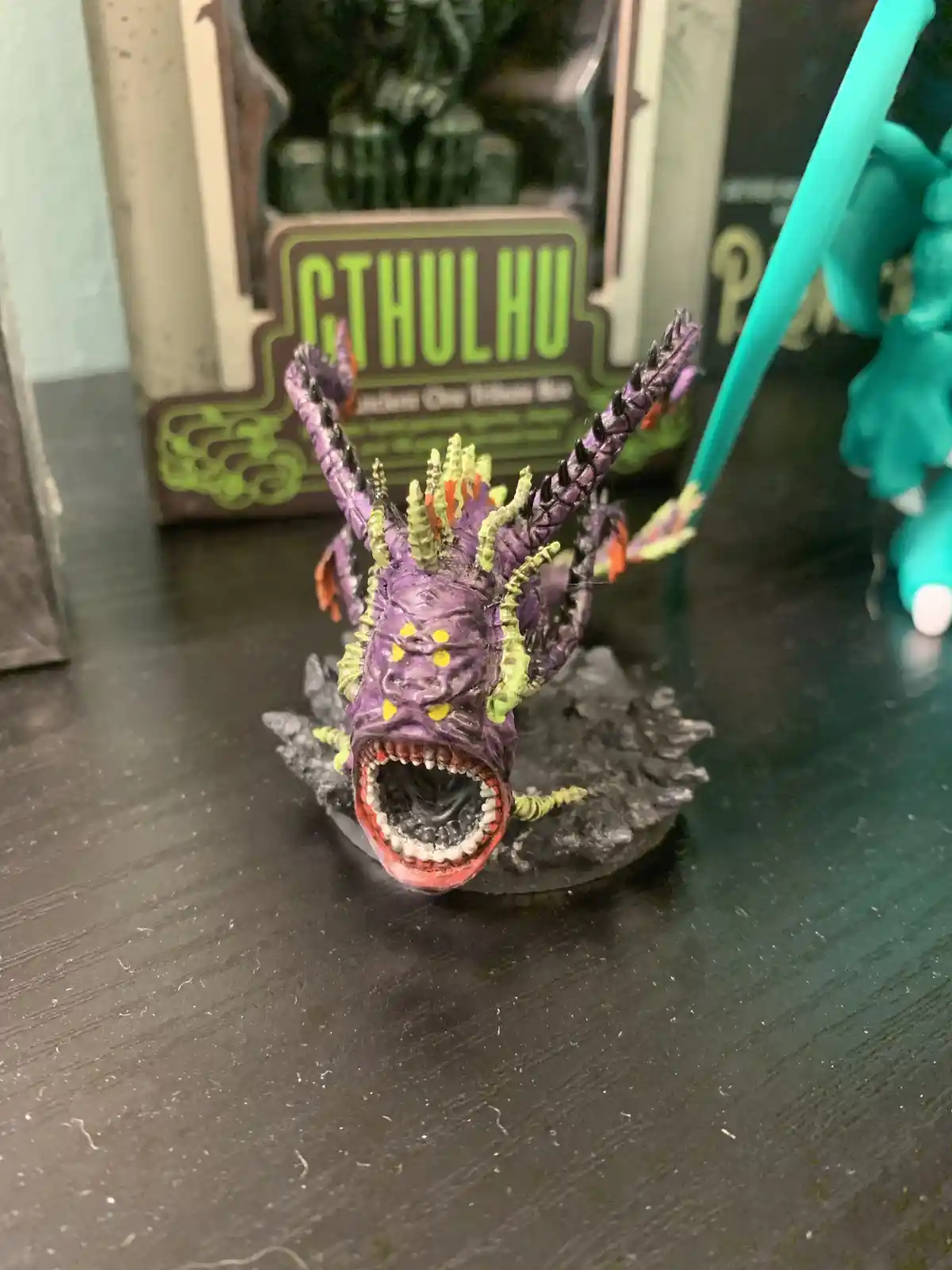 A small, purple, spiky Cthulhu figurine with a wide-open mouth and yellow eyes sits on a dark surface. Behind it, a box…