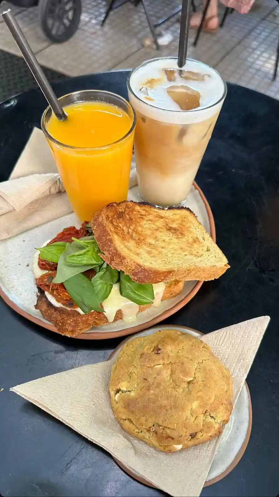 A table with a sandwich topped with basil and sun-dried tomatoes, a glass of orange juice, an iced coffee, and a large…