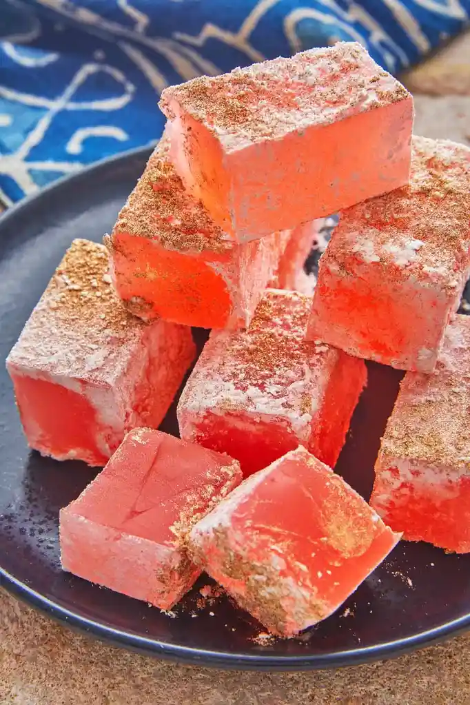 A pile of bright pink, cube-shaped confections dusted with white powder and gold glitter sits on a dark plate. The image is…