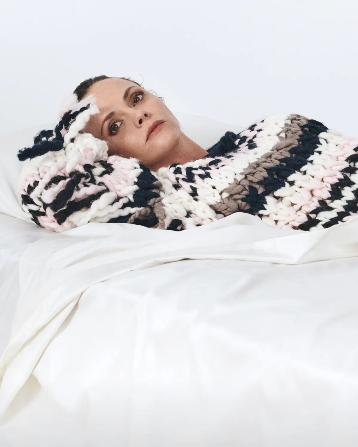 A woman lies in bed under a thick, chunky knit blanket with stripes of white, pink, black, and gray, looking upward with a…