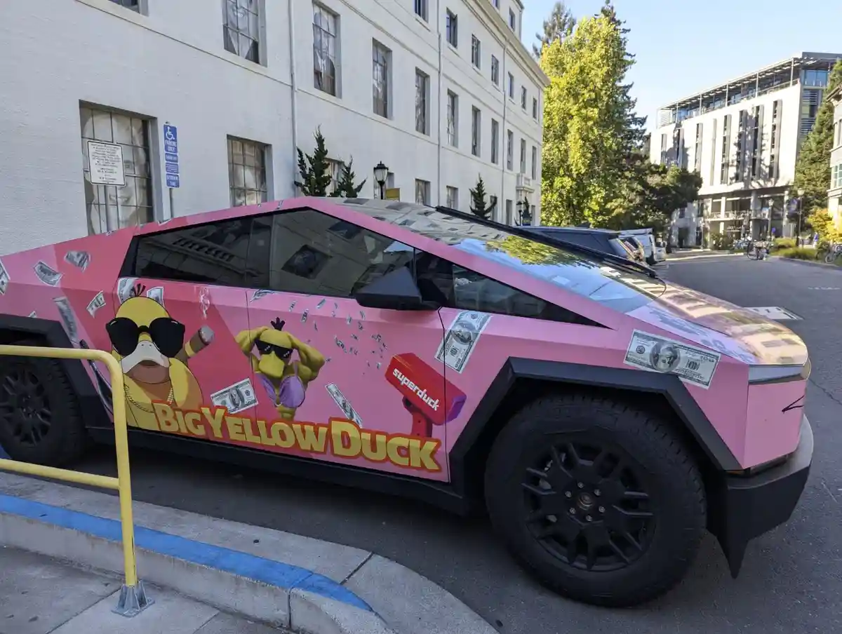 A pink Tesla Cybertruck is parked on a city street, covered in decals of cartoonish yellow ducks wearing sunglasses…