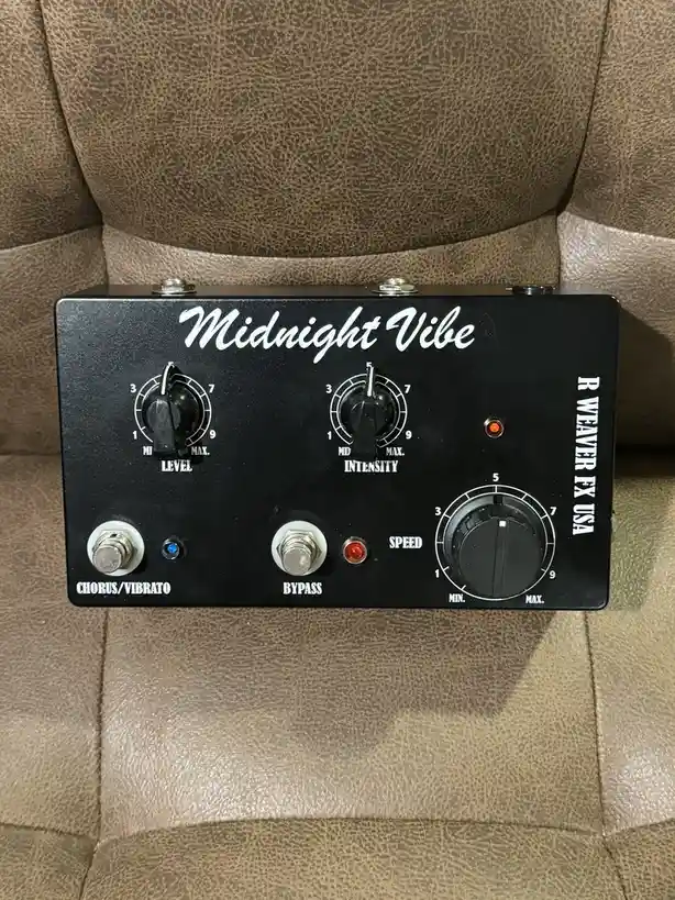 A black guitar effects pedal labeled 'Midnight Vibe' by R Weaver FX USA sits on a brown leather couch. It has three knobs…