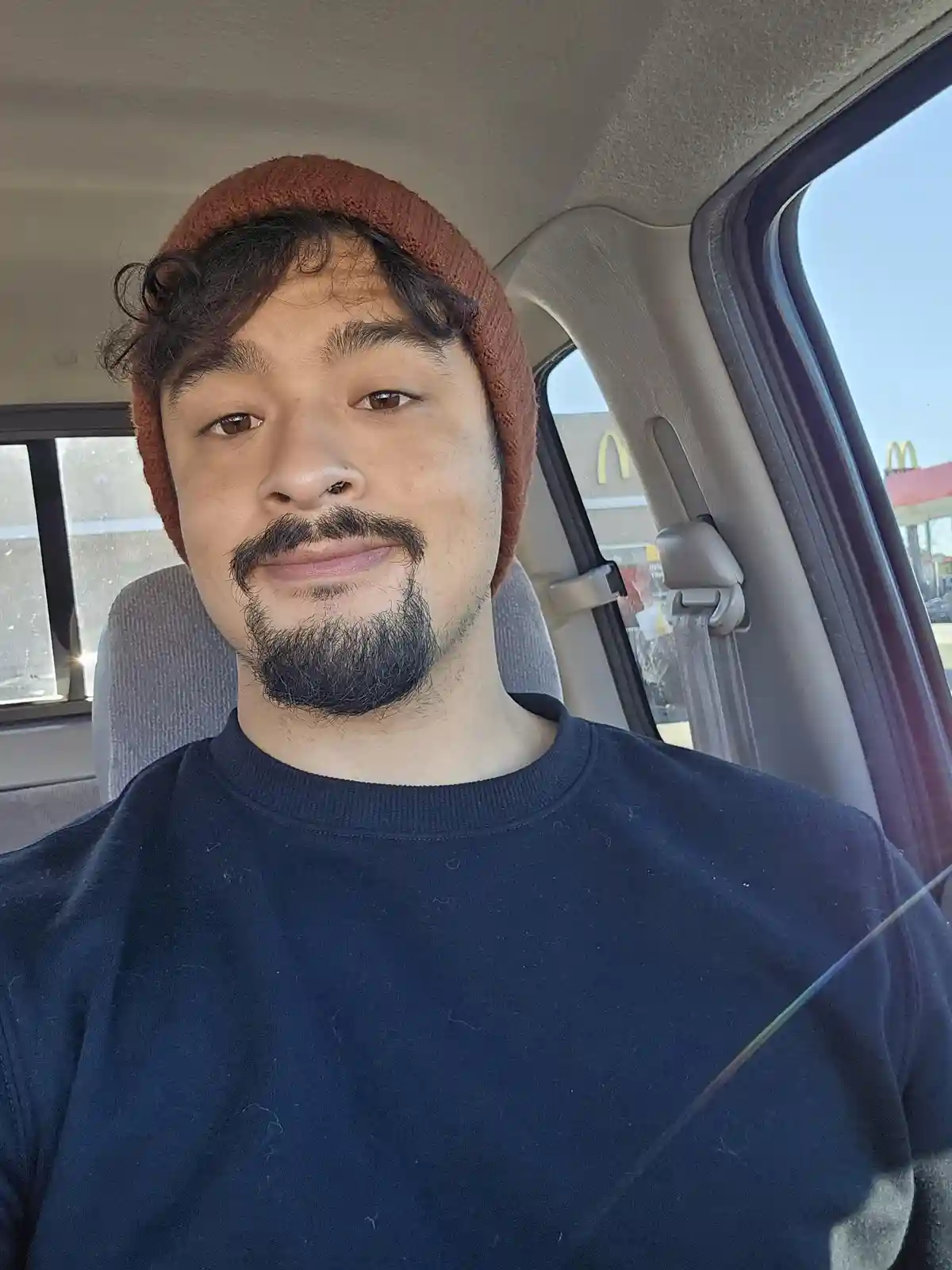 A man with a brown beanie and dark beard sits in a car, looking at the camera with a slight smirk. Through the car window, a…