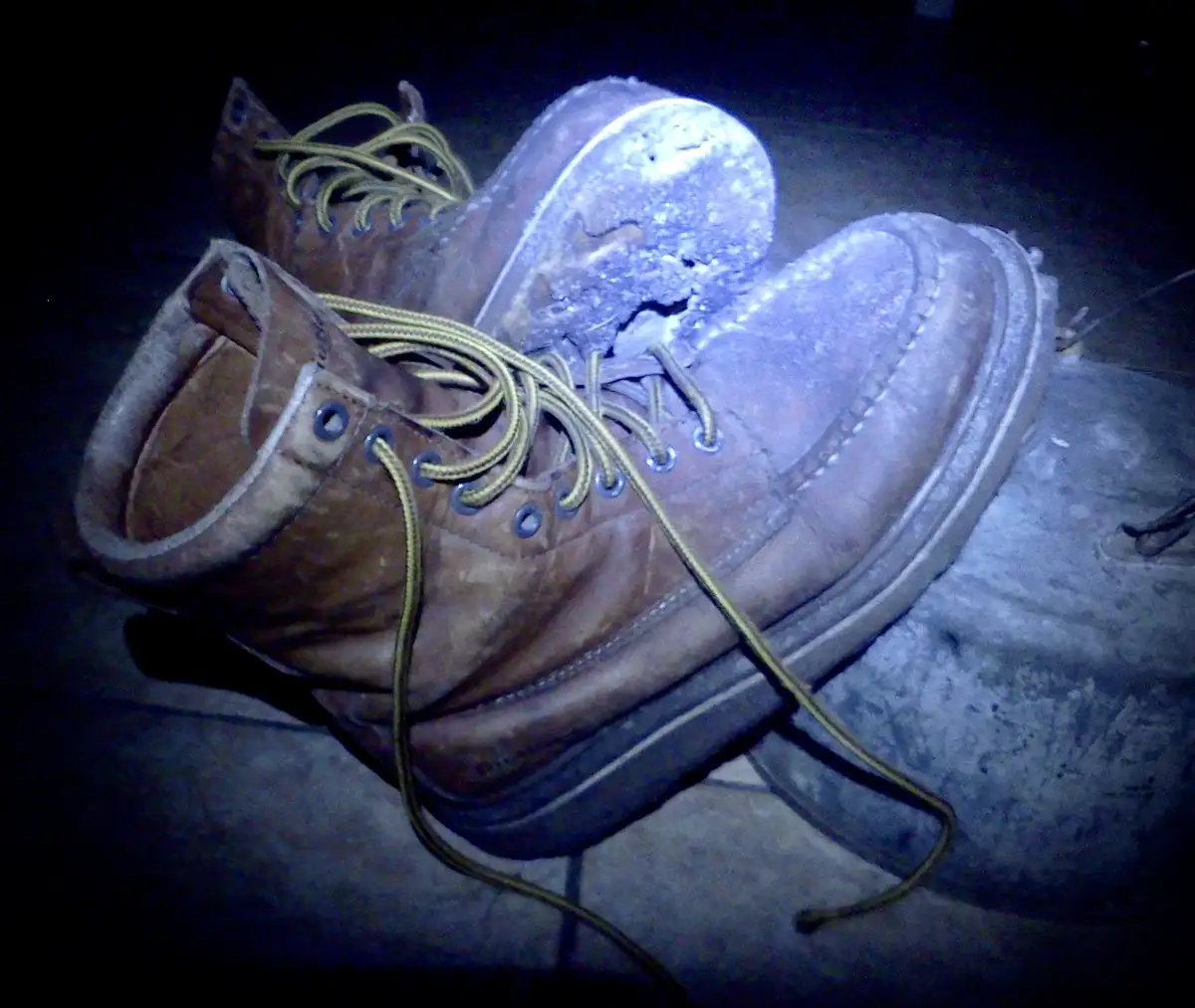 A pair of heavily worn, dirty brown work boots with frayed laces and a cracked, worn-out sole are shown in dim lighting. The…