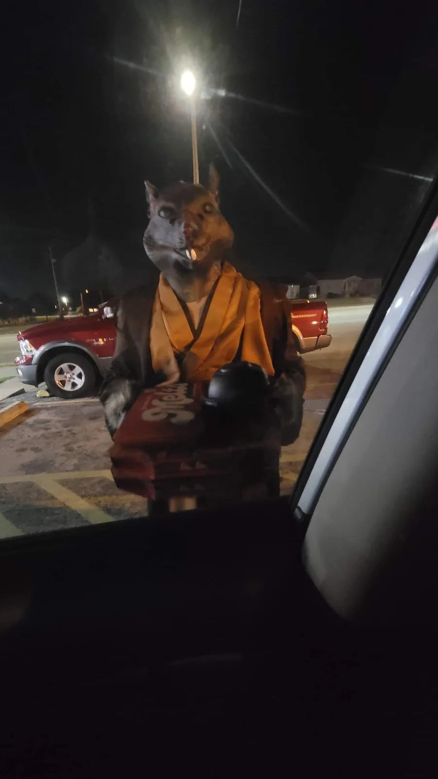 A person wearing a large, realistic rat head mask and an orange monk robe stands outside a car at night, holding a tray with…