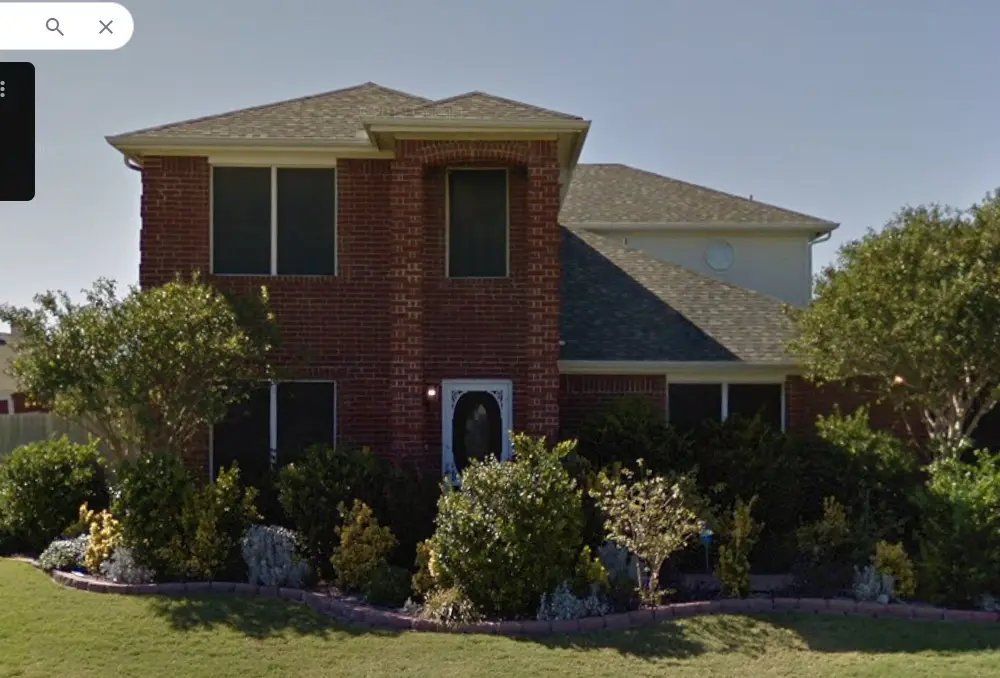 a google street view image of a two-story brick house with a white front door, surrounded by green bushes and trees under a…