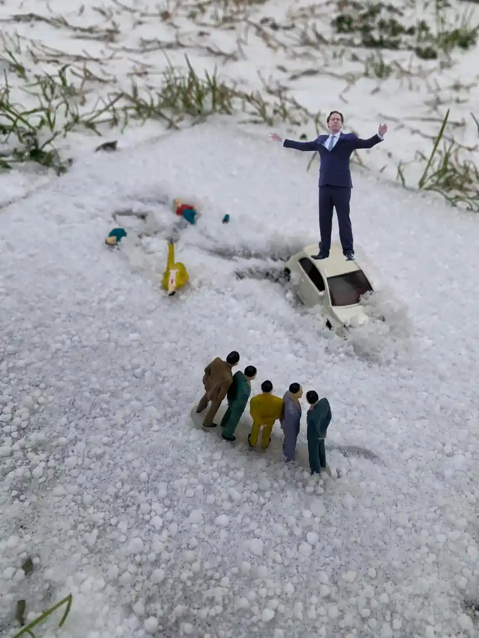 A miniature figurine of a man in a suit stands atop a toy car buried in snow, arms outstretched, while other miniature…