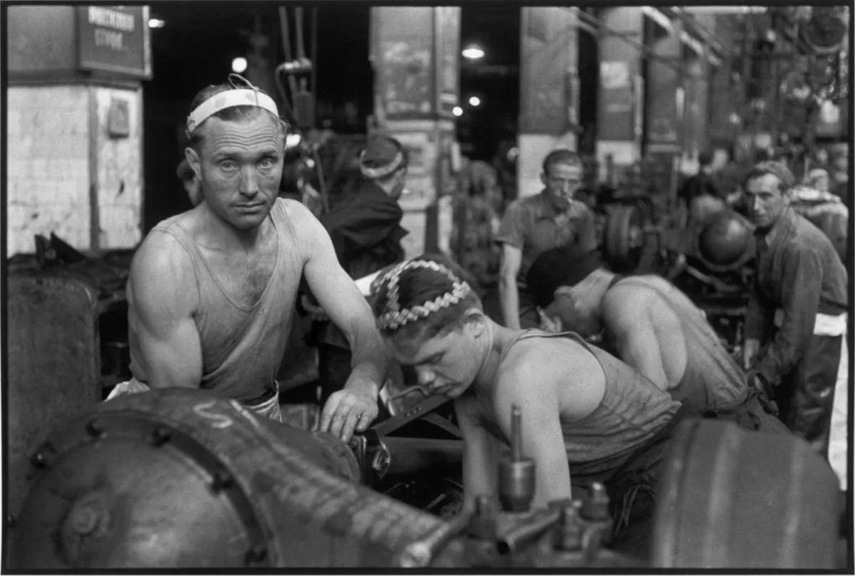 A black-and-white historical photo shows several men in a factory or workshop, wearing headbands and tank tops, working on…