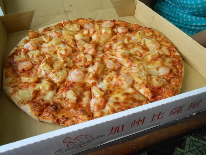 A whole Hawaiian pizza with shrimp and pineapple toppings sits inside an open cardboard pizza box. The box's side features…