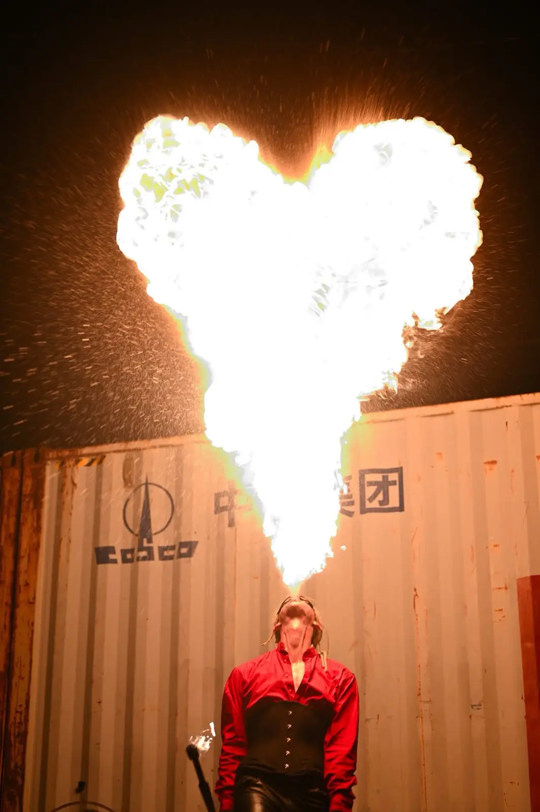 A person in a red shirt and black corset stands in front of a shipping container at night, exhaling a large, bright…