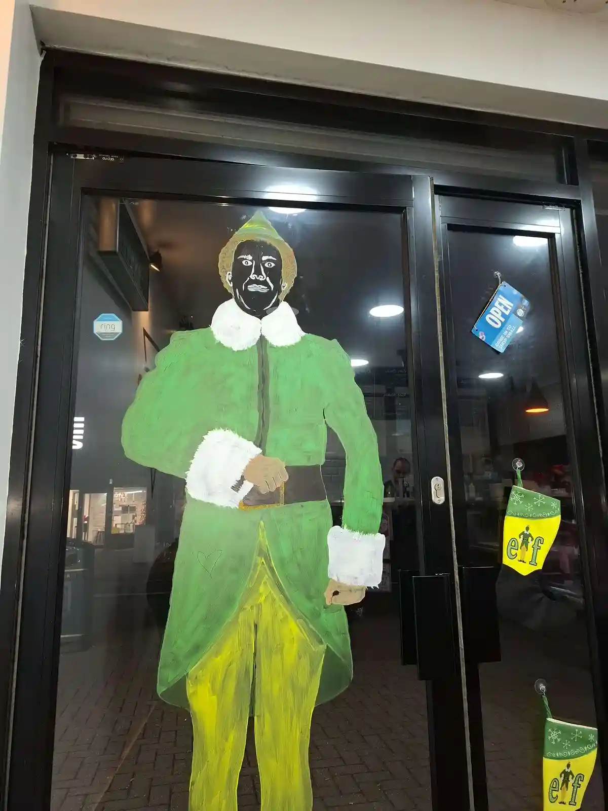 A hand-painted figure of an elf in a green coat and yellow pants stands on a glass storefront door, with a blue 'OPEN' sign…