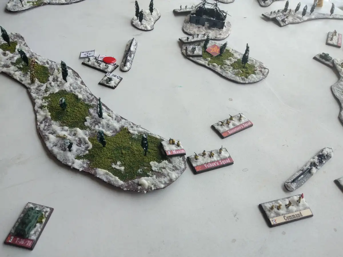 A tabletop wargame setup depicts a snowy battlefield with miniature figures and terrain pieces, including squad bases…