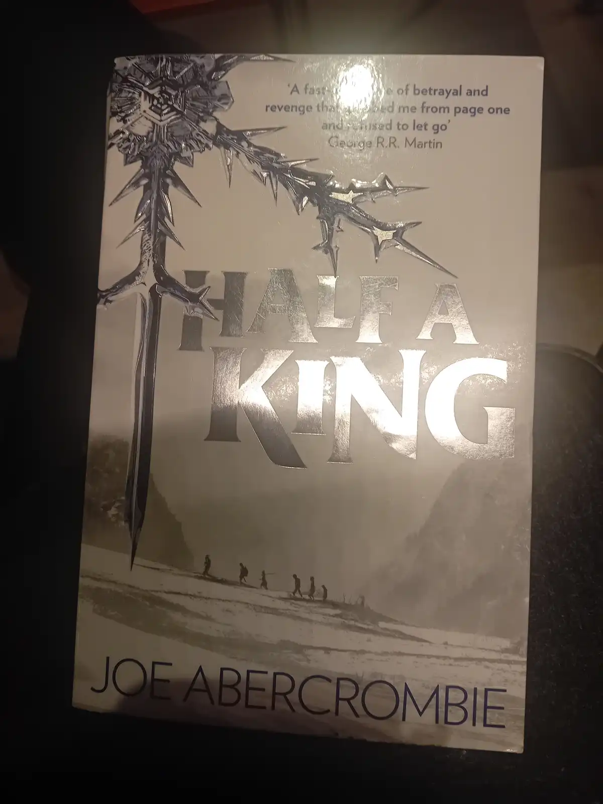 The image shows the cover of the book 'Half a King' by Joe Abercrombie. The cover features a large, ornate sword with a…
