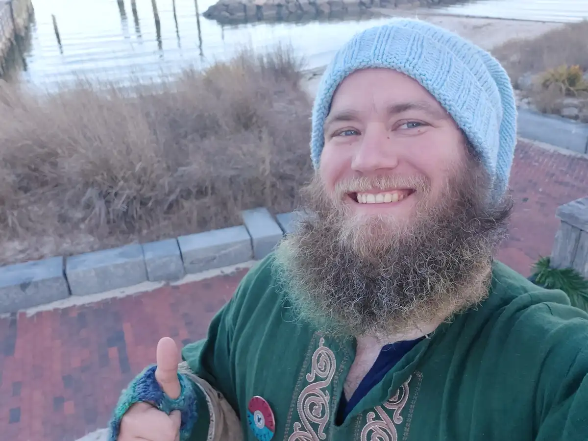 A man with a long beard and a light blue knit beanie smiles at the camera while giving a thumbs-up. He wears a green robe…