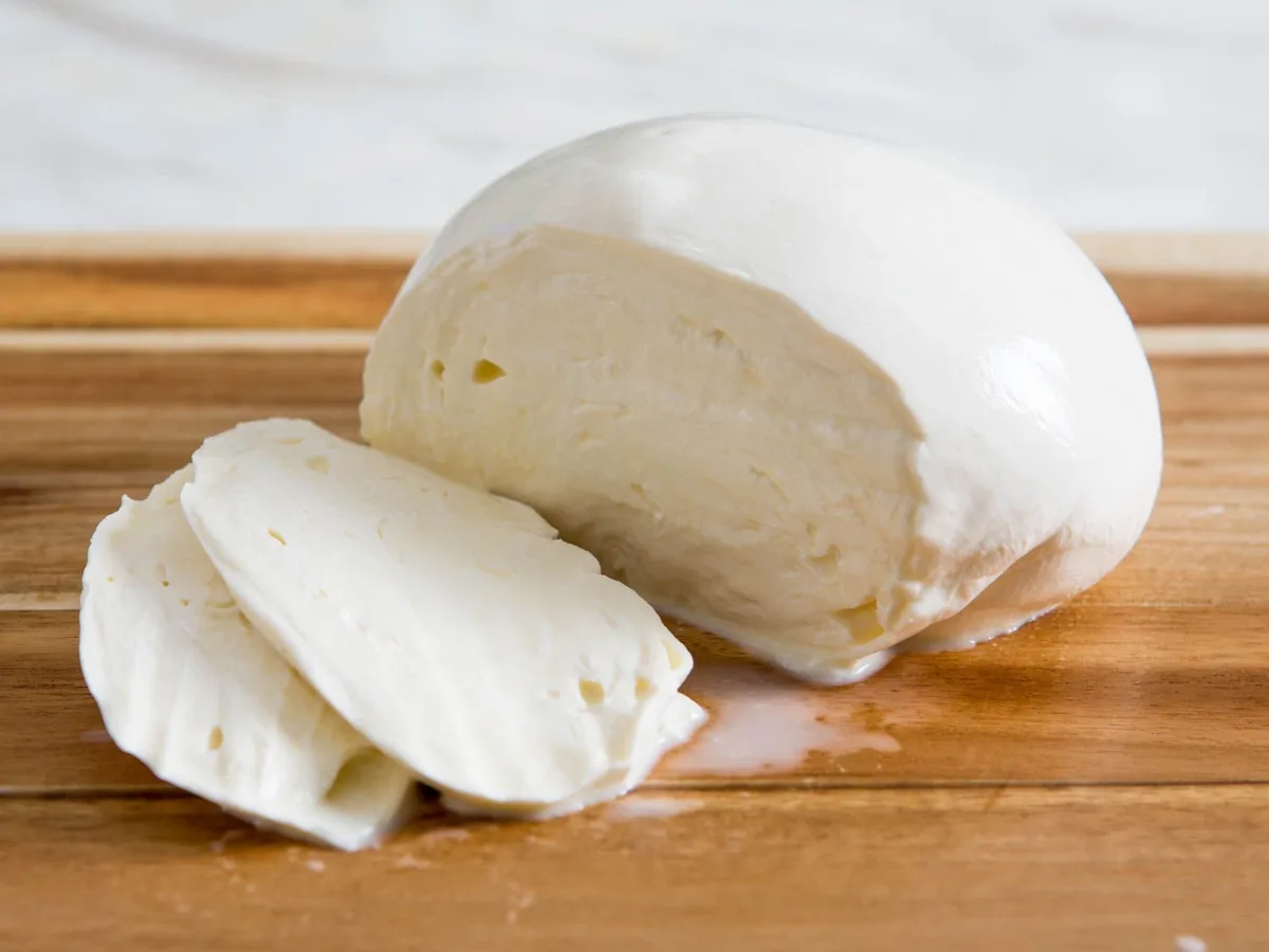 A close-up photo shows a large, round ball of fresh white mozzarella cheese sitting on a wooden cutting board, with a few…