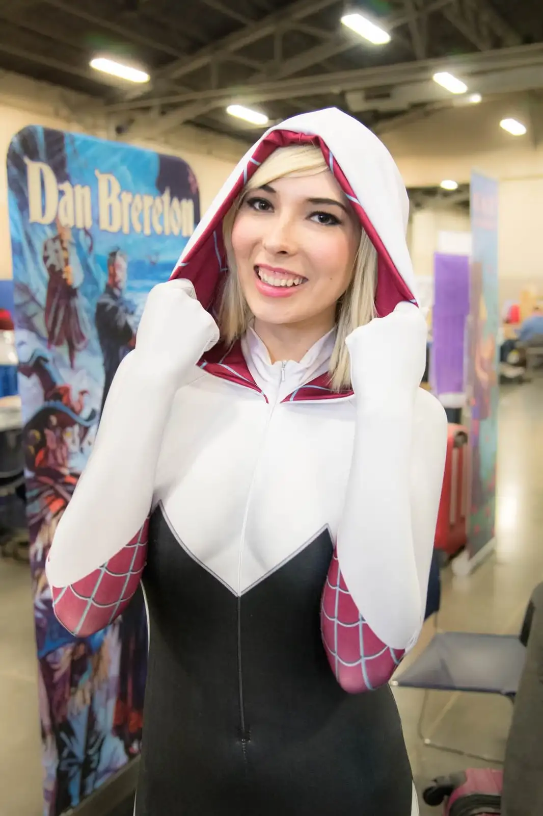A woman in a white, black, and red Spider-Gwen cosplay costume poses with her fists up at a convention, smiling. Behind her…
