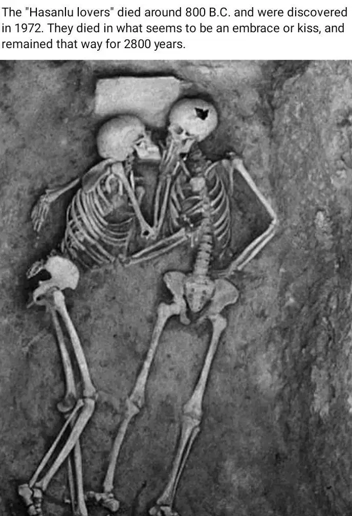 A black-and-white photograph shows two human skeletons lying face-to-face in an embrace or kiss, buried in earth. The text…