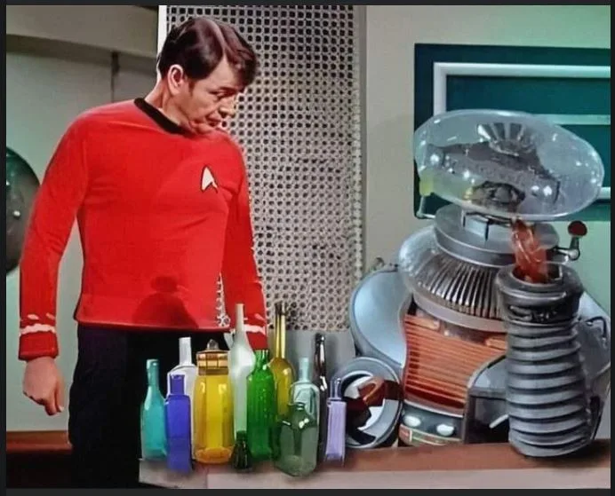 A man in a red Star Trek uniform stands beside a cluttered table with colorful glass bottles and a large, complex, silver…