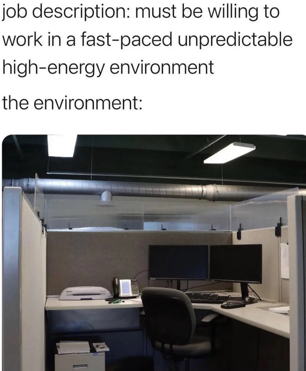 An office cubicle with two black monitors, a keyboard, a phone, a printer, and a chair, all under fluorescent lights and a…