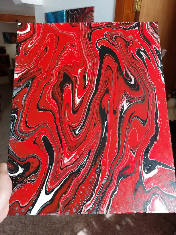A person holds up a large, square canvas featuring a vibrant, abstract painting with swirling patterns of red, black, and…