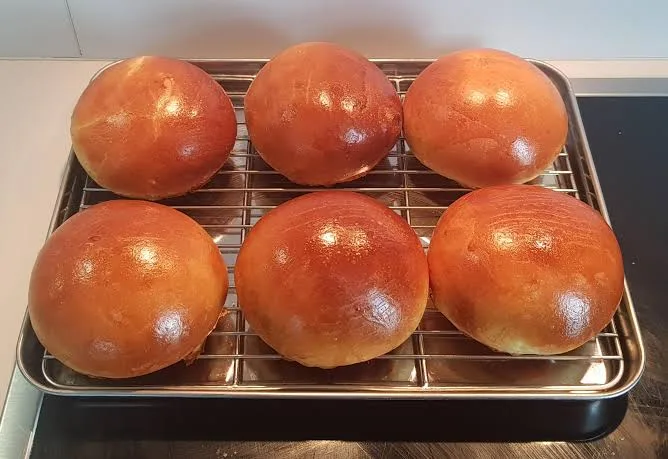 Six perfectly round, golden-brown baked buns sit evenly spaced on a metal wire cooling rack, which rests on a dark baking…