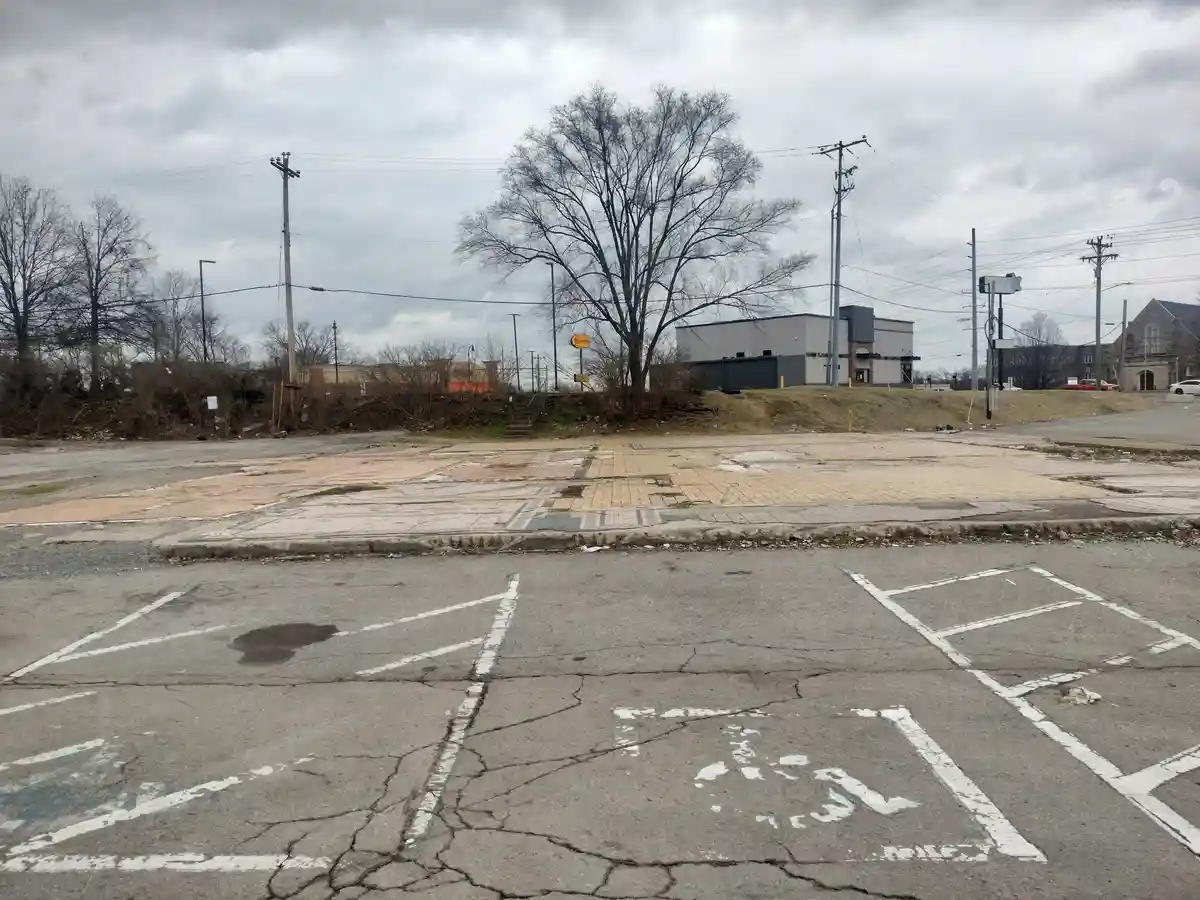 A cracked, empty asphalt parking lot with faded white lines and a broken "police" marking, under an overcast sky, with a…