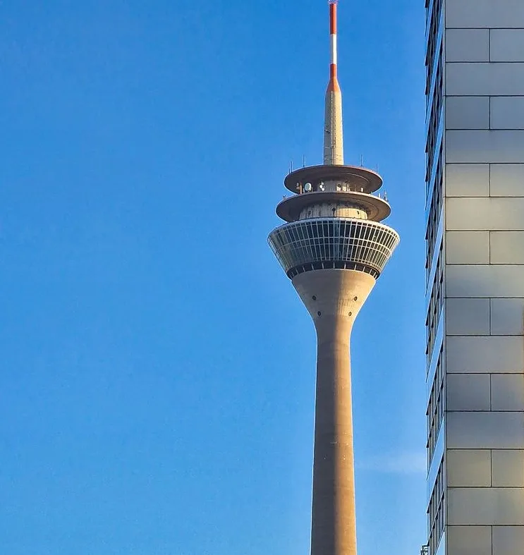A tall, slender observation tower with a circular viewing platform and red-and-white striped antenna against a clear blue…