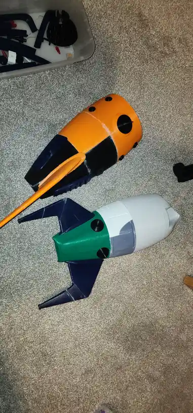 Two handmade, 3D-printed model spacecraft lie on a carpeted floor: one orange and black with a long orange tail, the other…