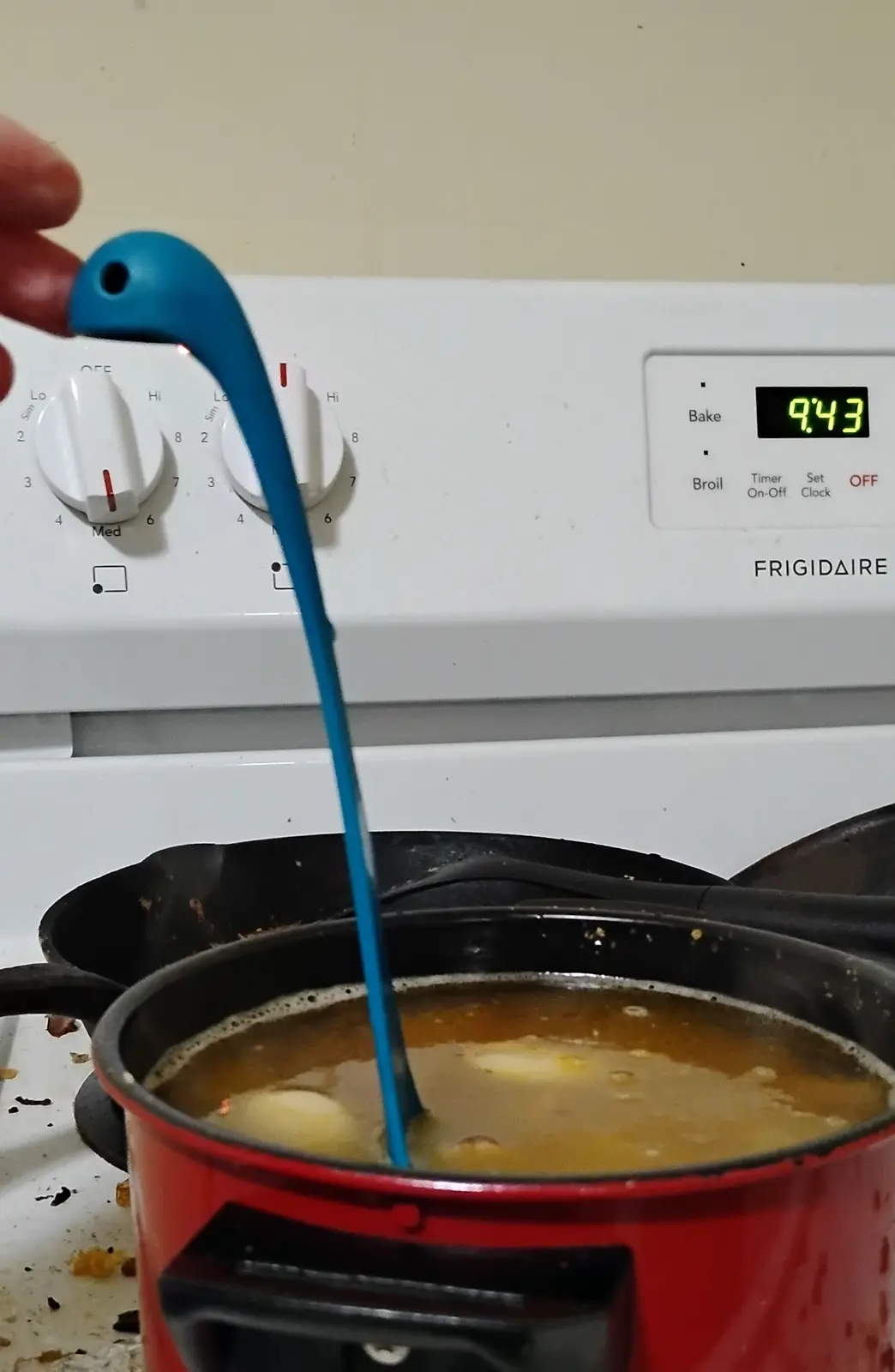A hand holds a blue spoon over a red pot of simmering broth with eggs, positioned above a white Frigidaire stove with a…