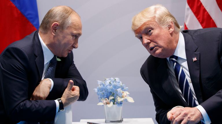 Two men in suits sit across a table from each other, facing one another with serious expressions. Behind them are national…