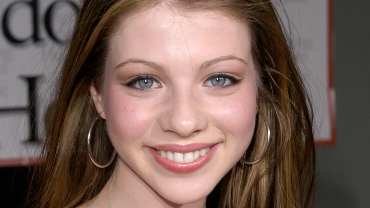 A close-up photo of a young woman with light brown hair, blue eyes, and large hoop earrings, smiling at the camera. The…