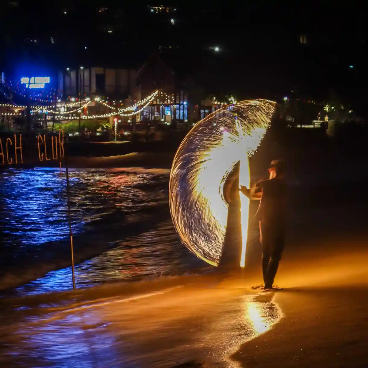 A person stands on a beach at night, spinning a flaming torch to create a bright, circular light trail. Behind them, the…