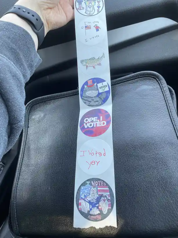 A hand holds a strip of six circular 'I Voted' stickers over a black car seat. The stickers feature various designs…