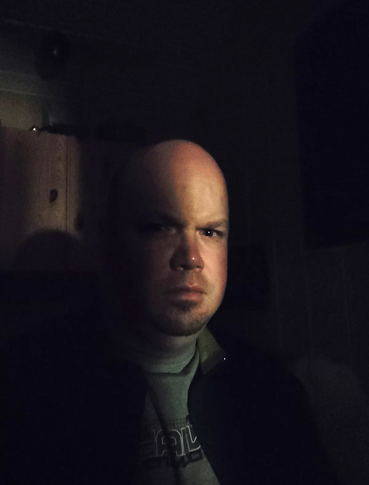 A bald man with a neutral to slightly stern expression stares directly at the camera in a dimly lit room. He wears a dark…