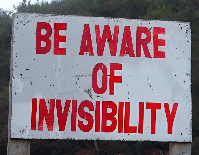 A weathered white sign with bold red text reads 'BE AWARE OF INVISIBILITY' against a backdrop of trees. The sign shows…