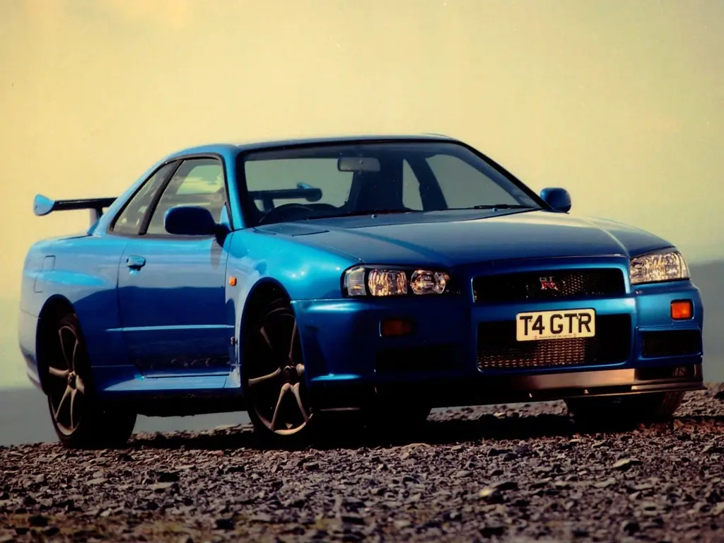 A blue Nissan Skyline R34 GTR is parked on a gravel surface with a hazy sky background. The car's front grille displays a…
