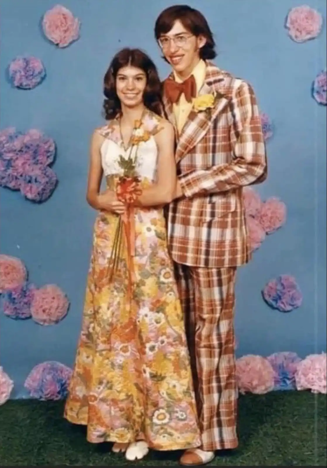 A vintage photo of a young couple posing for a portrait against a blue backdrop decorated with pink and purple paper…