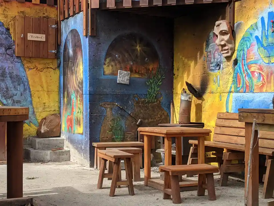 An outdoor seating area with wooden tables and benches sits before a colorful, weathered wall covered in murals and art. The…