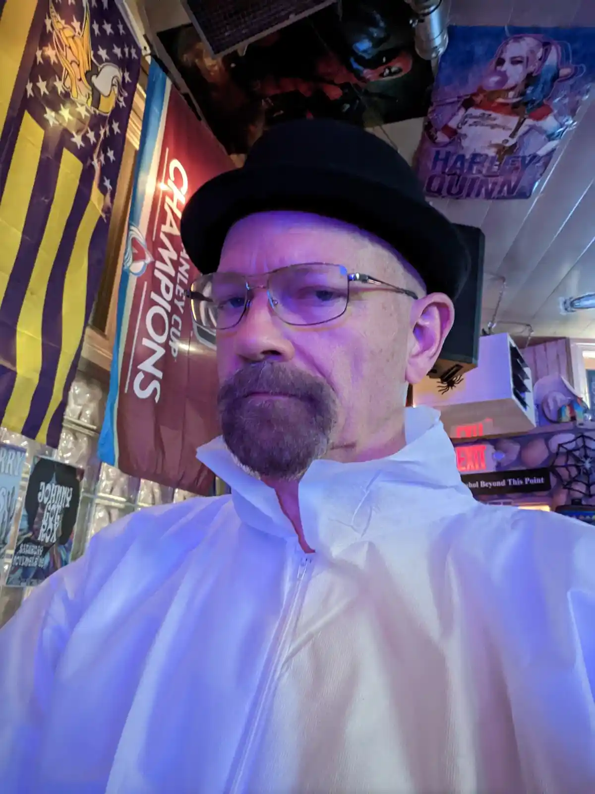 A man wearing a black bowler hat, glasses, and a white zip-up jacket resembling a lab coat looks directly at the camera with…
