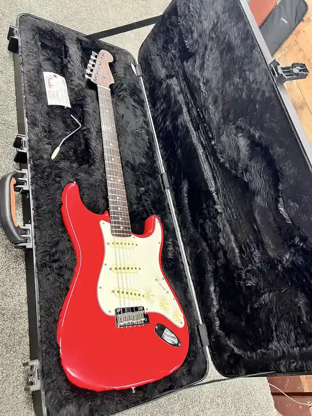 A bright red Fender Stratocaster electric guitar rests inside an open, black, plush-lined hard case, with a small white…