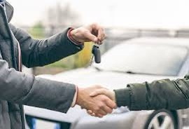 Two people are shaking hands while one person hands over a set of car keys to the other. The background shows a silver car…