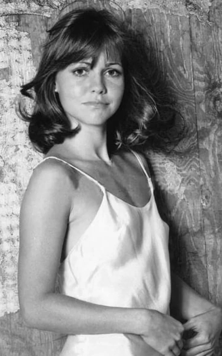 A black-and-white photo of a woman with shoulder-length wavy hair and bangs, wearing a white camisole, standing against a…