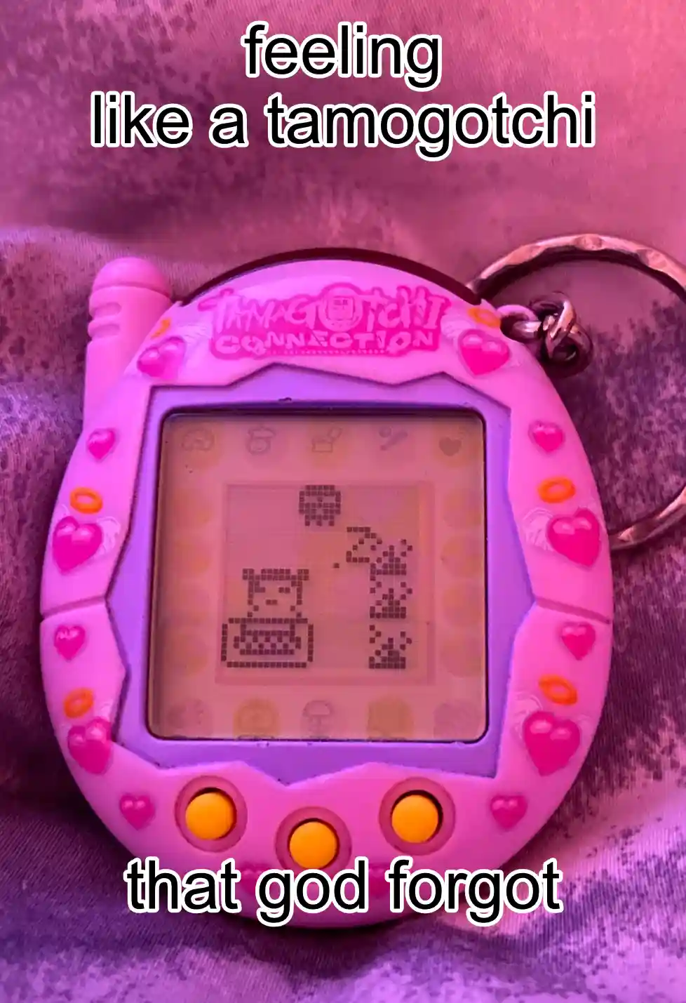 A pink Tamagotchi Connection handheld game device with a pixelated screen showing a sad-looking creature, overlaid with the…