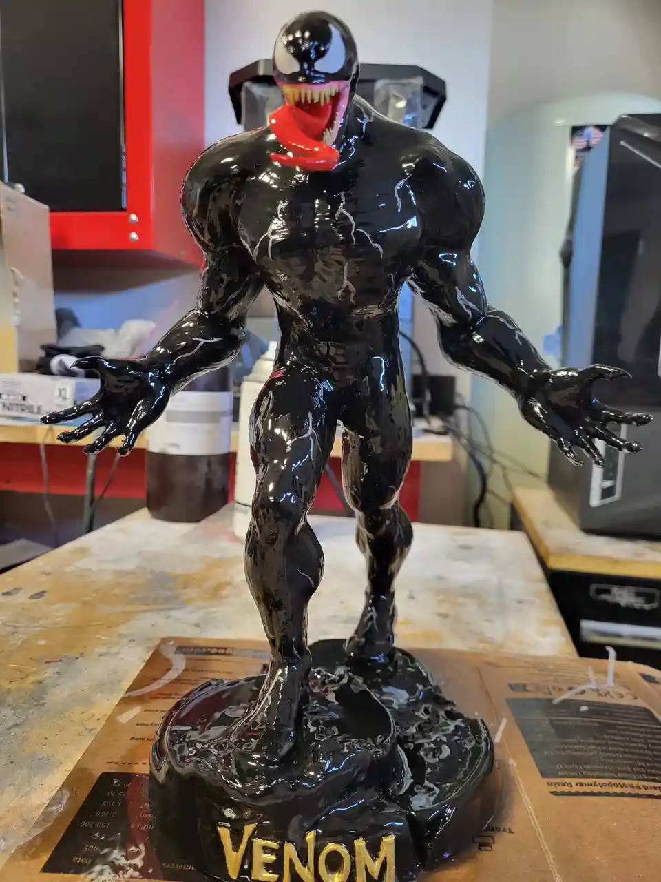 A glossy black Venom statue with a wide-open mouth and red tongue stands on a messy workbench, its base inscribed with…