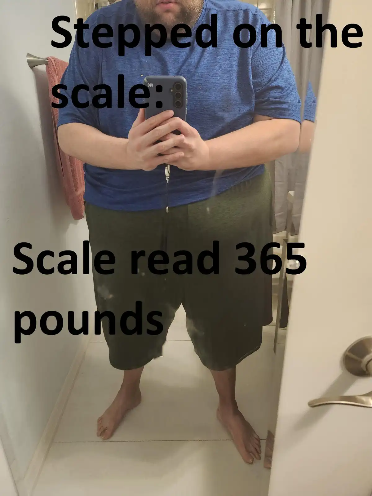 A man stands barefoot in a bathroom mirror selfie, wearing a blue t-shirt and green shorts, holding a phone. Text overlaid…