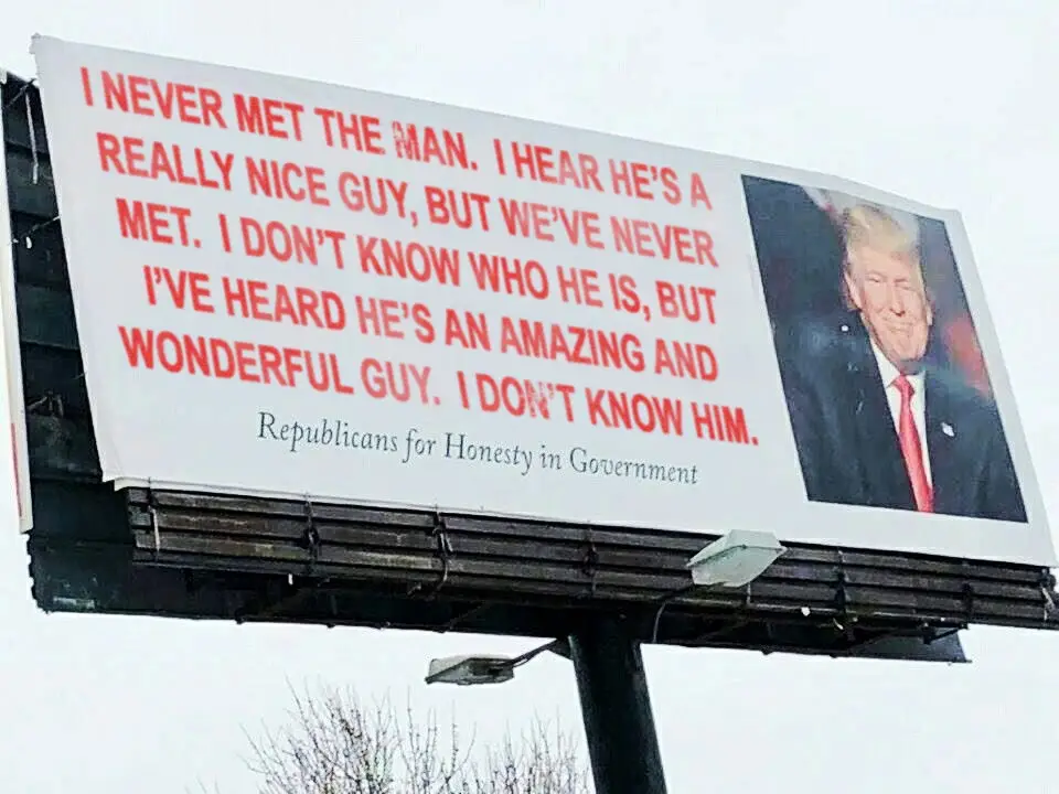 A large outdoor billboard displays a photo of Donald Trump alongside red text that repeatedly states he has never been met…